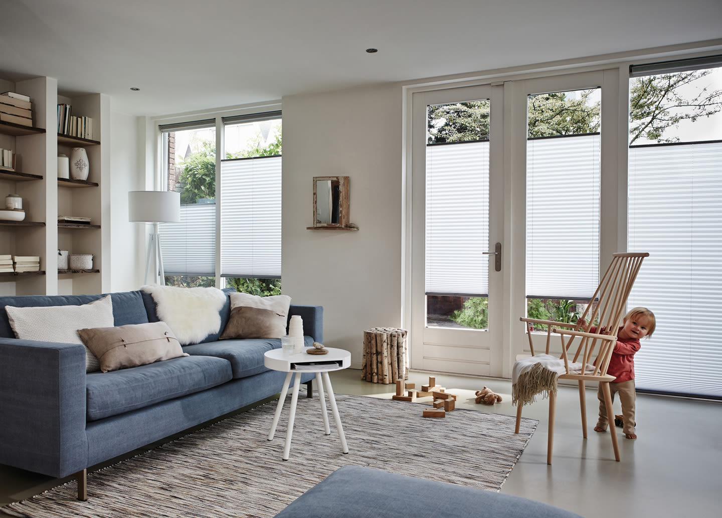 Lyse stue med blå sofa, hvitt rundt bord, trestol med pledd og hvite foldegardiner