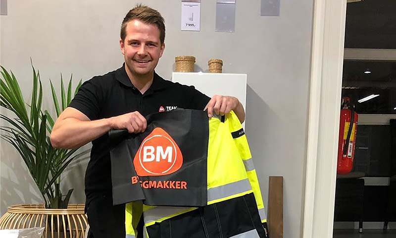 Mann i svart t-skjorte holder svart handlenett med oransje BM-logo og gul refleksjakke.
