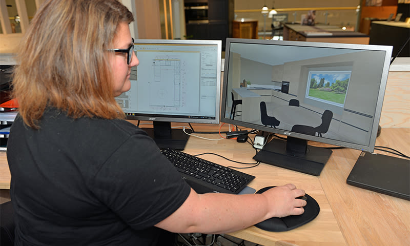 Person i svart t-skjorte ved trebord med to skjermer, én viser 2D-plan, én 3D kjøkkeninteriør.