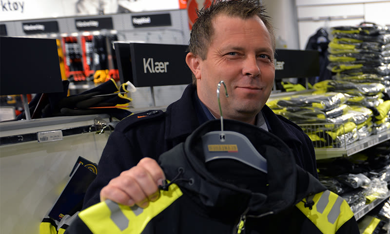 Person holder svart-gul sikkerhetsjakke i butikkgang med hyller fulle av klær.