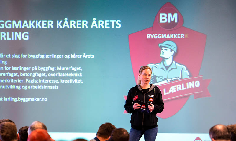 Person på scenen holder premie foran blåhvit bakgrunn med rødt Byggmakker-logo og publikums hoder