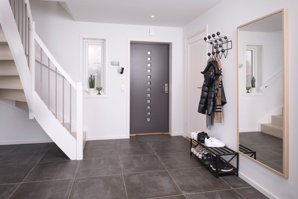 Hvit entrée med mørkgrå fliser, svart dør, jakker på veggknagger, skohylle og speil.