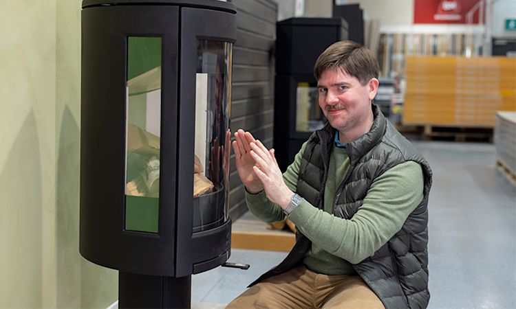 Mann i grønn genser varmer hendene ved svart ovn med vedfyrt flamme i byggevarebutikk.
