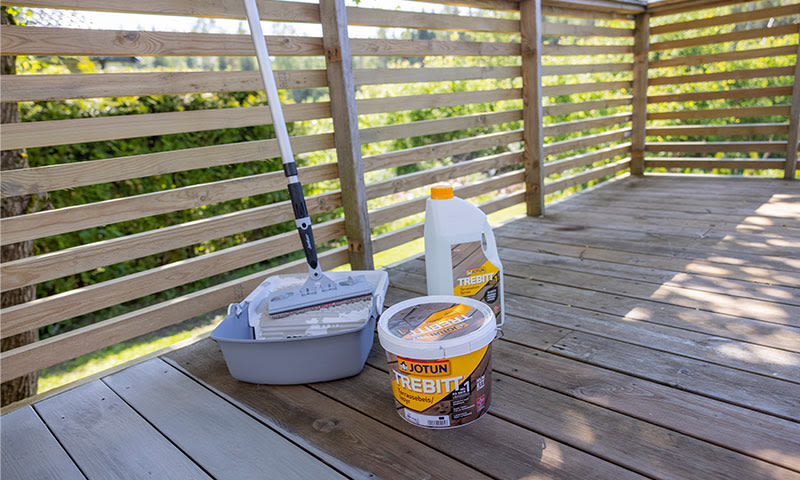 Gul Trebitt-malingsbøtte, hvit kanne og malerulle på solrik treterrasse med gjerde.