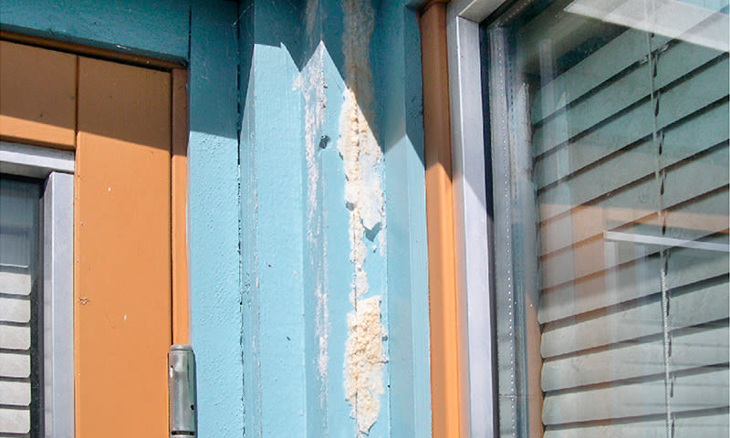 Blå søyle med avflasset maling og hvitt isolasjonsskum mellom oransje vinduskarmer.