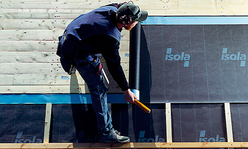 Bygningsarbeider i mørkt arbeidstøy ruller ut svart Isola-takmembran på lyst treverk