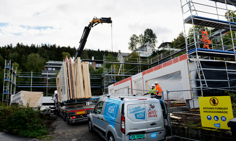 Byggeplass med stillas og hvite moduler, kran løfter trepaneler, oransjekledde arbeidere