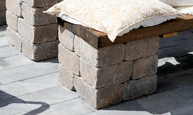 Utebenk av grå betongblokker og treplate med hvit-gule puter på grå heller.