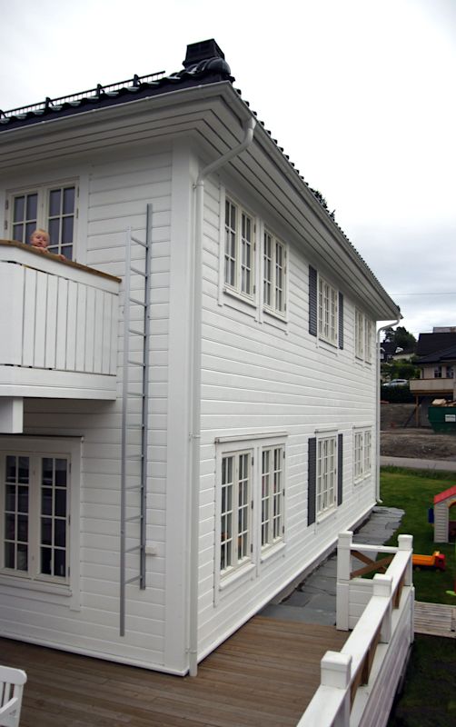 Hvitt toetasjes trehus med svart tak, flere vinduer, balkong, stige og terrasse.