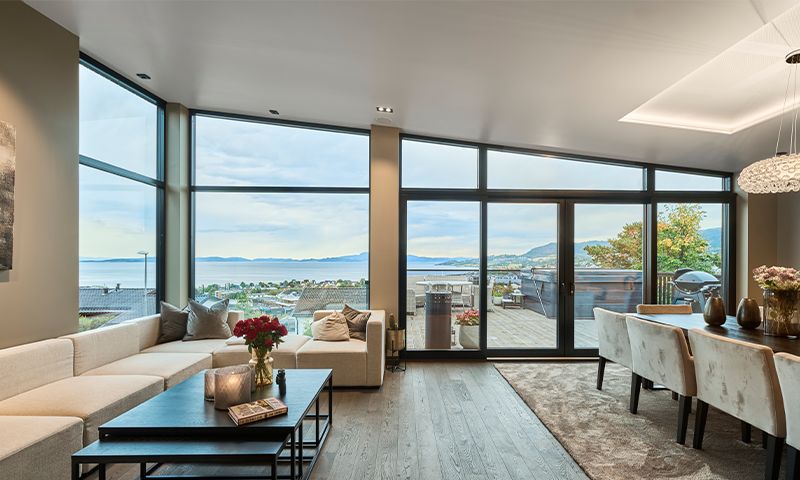 Moderne lys stue med store vinduer mot fjord, beige sofa, tremøbler og spiseplass.