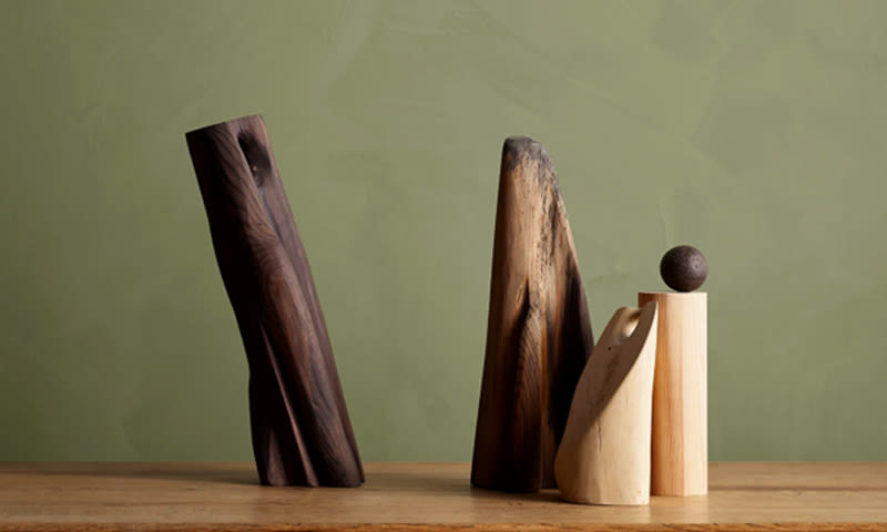 Fire abstrakte treskulpturer i brunt med en kule, på bord foran olivengrønn vegg.