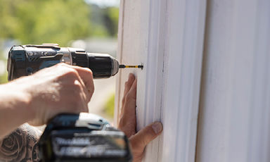 Nærbilde av en hånd som bruker svart batteridrevet drill til å skru en skrue inn i hvit trepanel.