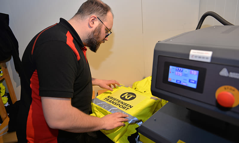 Mann trykker gul refleksjakke med «Kristensen Transport»-logo på pressmaskin.