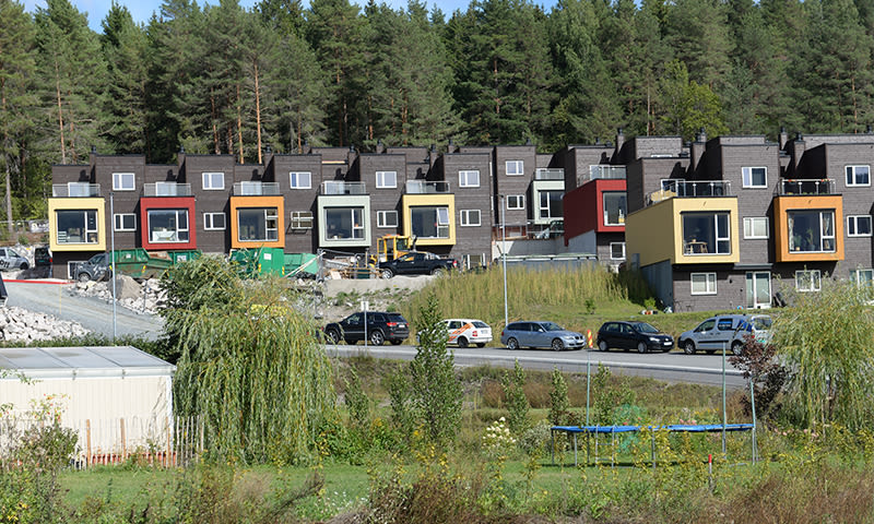 Moderne rekkehus med fargerike kasseutstikkere ved skog, byggeplass og biler foran.