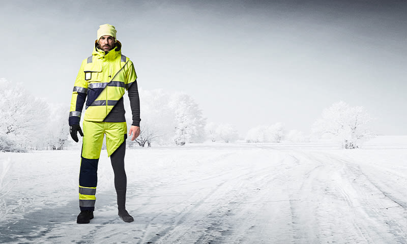 Person i neongul refleksjakke og -bukse på snødekt vei foran hvite trær.