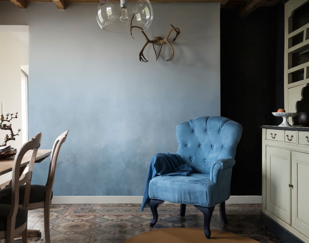 Lyst rom med blå fløyelslenestol, tremøbler, vegghengt gevir og glasslampe.