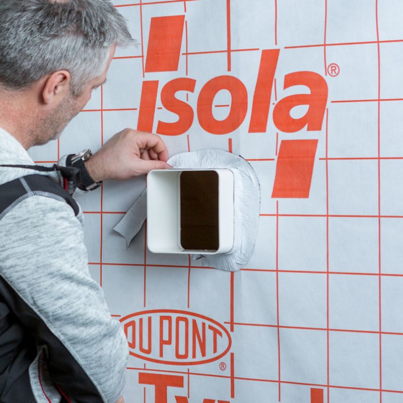 Hånd monterer hvit ventilkanal gjennom hvit duk med rødt rutenett, Isola- og DuPont-logoer.