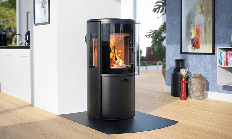 Svart sylinderformet vedovn med flammende ild i lyst rom med blå vegg og lyst tregulv.