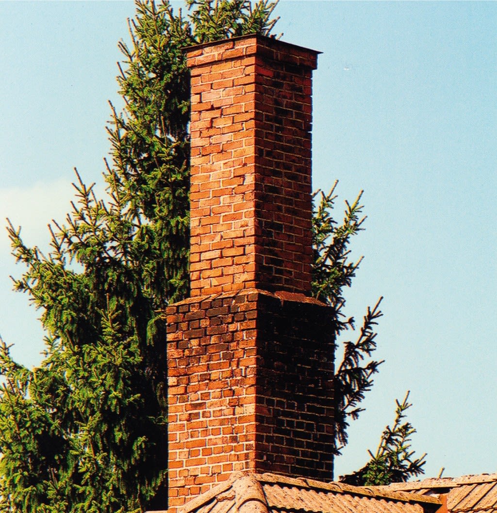 Rød mursteinsskorstein på tak med grantrær og blå himmel i bakgrunnen.