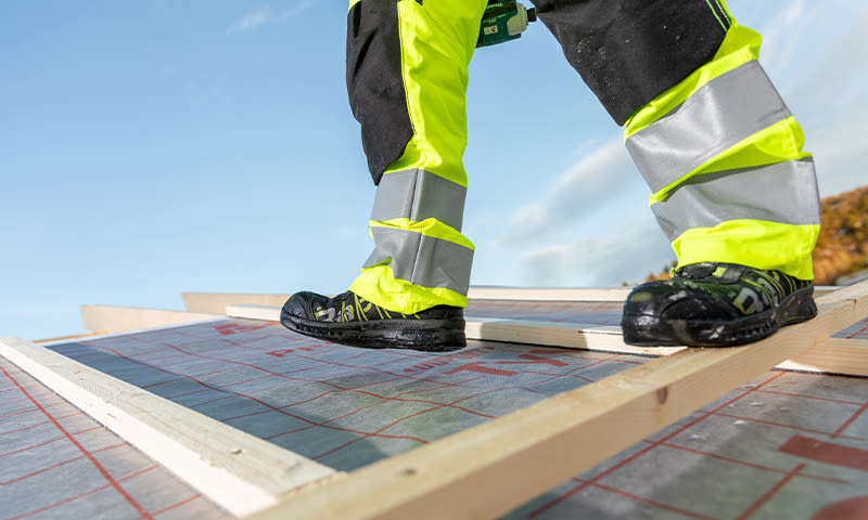 Nærbilde av arbeider i neongul refleksbukse og vernesko på taklekter.
