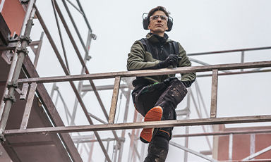 Byggearbeider i grønn jakke med hjelm og hørselsvern på metallstillas under grå himmel