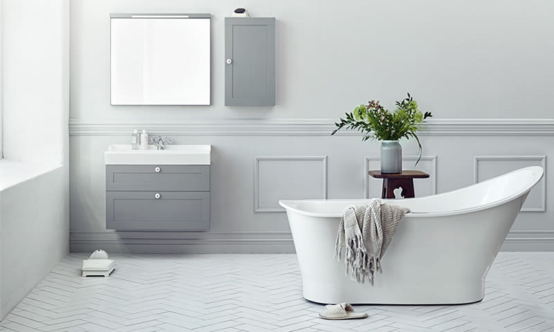 Lyst grå-hvitt baderom med frittstående badekar, grått håndkle, speil, vask og grønn plante.