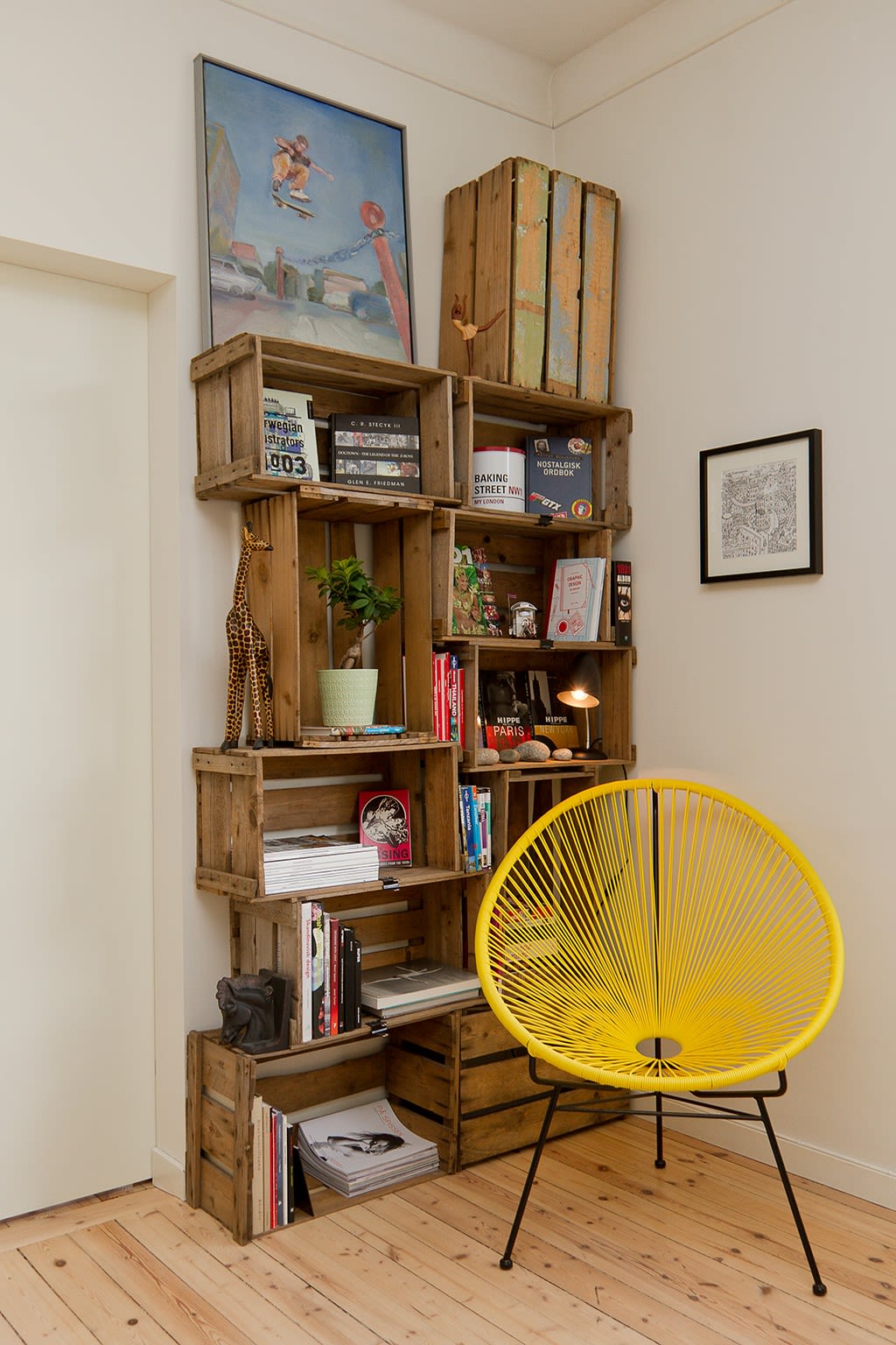 Hjørnehylle av trekasser med bøker, plante og maleri, foran gul flettestol på tregulv.