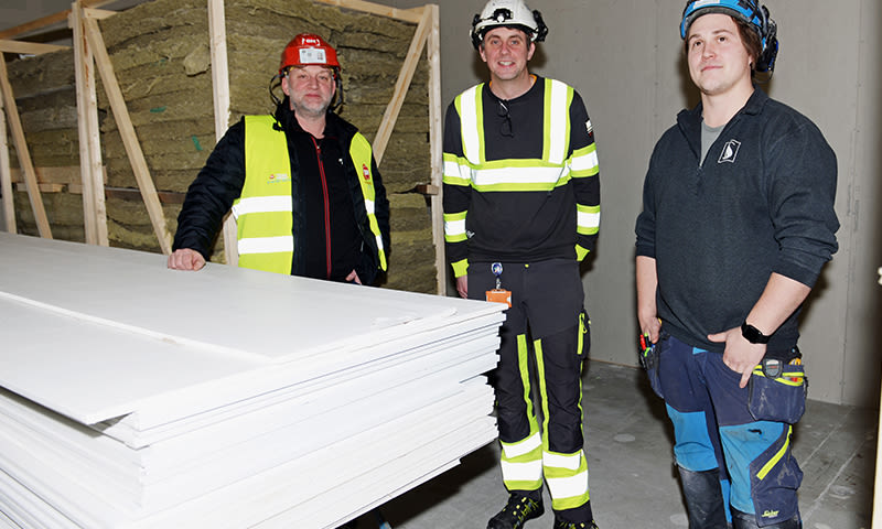 Tre byggearbeidere i hjelmer (rød, hvit, blå) og gule vester ved stablet hvit gipsplatebunke.