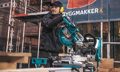 Arbeider i sort med gult hørselsvern bruker turkis kappsag foran rødt Byggmakker-banner.