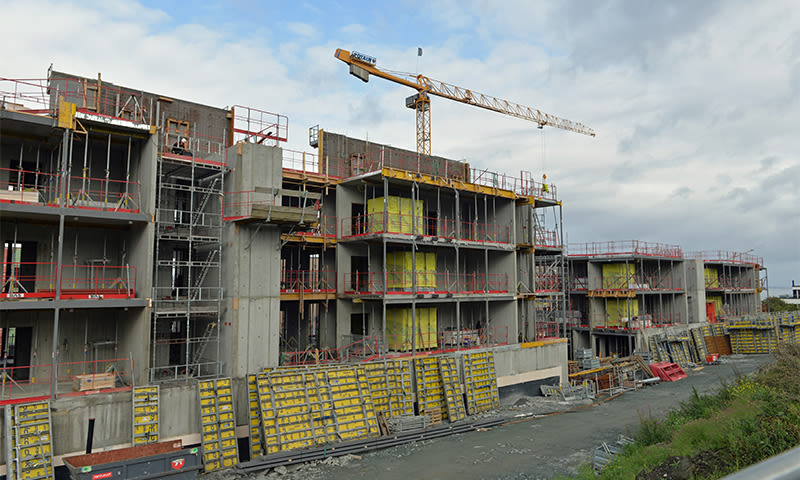 Etasjers betongbygg under oppføring, gule forskalingsplater, rødt rekkverk og kran mot skyet himmel