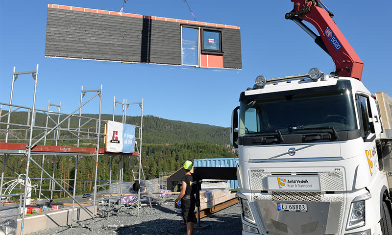 Hvit kranbil løfter svart-oransje modul med vindu over stillas ved skogkledd byggeplass.