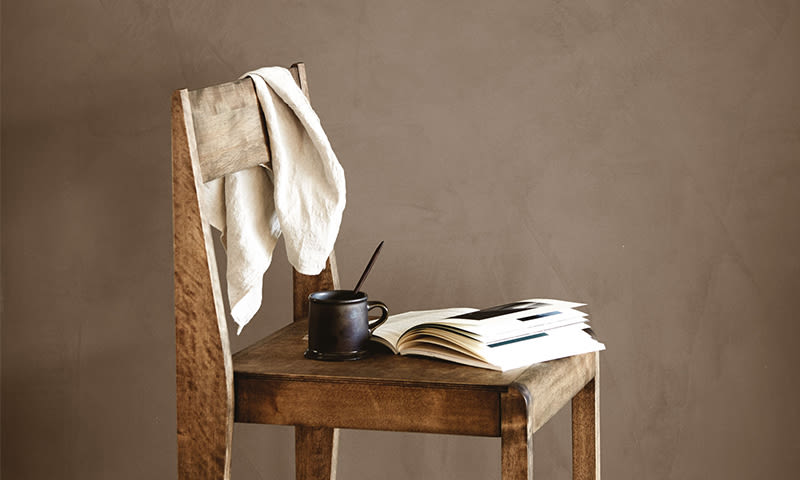 Trestol med hvit duk, svart kopp og åpen bok mot beige vegg.
