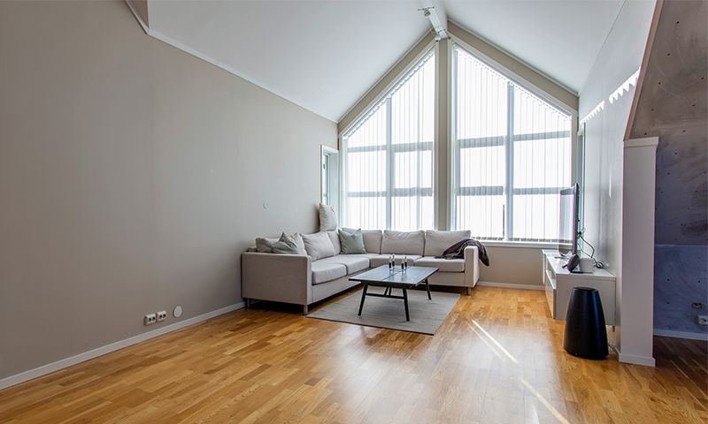 Lys stue med grå L-sofa, svart sofabord, store skråvinduer og lyst tregulv.