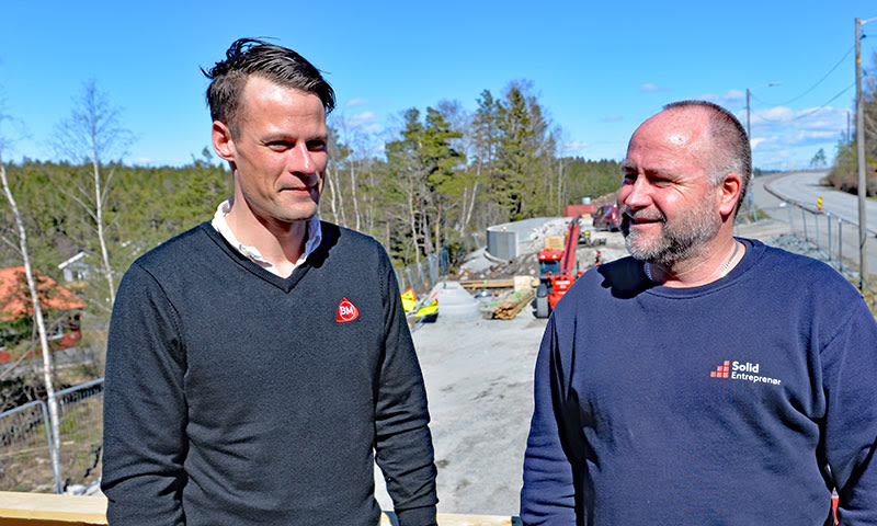To menn i mørke gensere på solfylt byggeplass ved vei, omgitt av skog og blå himmel.