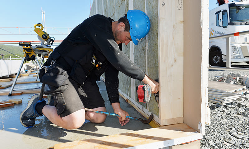 Bygningsarbeider i blå hjelm og svarte klær monterer trepanel ved byggeplass ute.