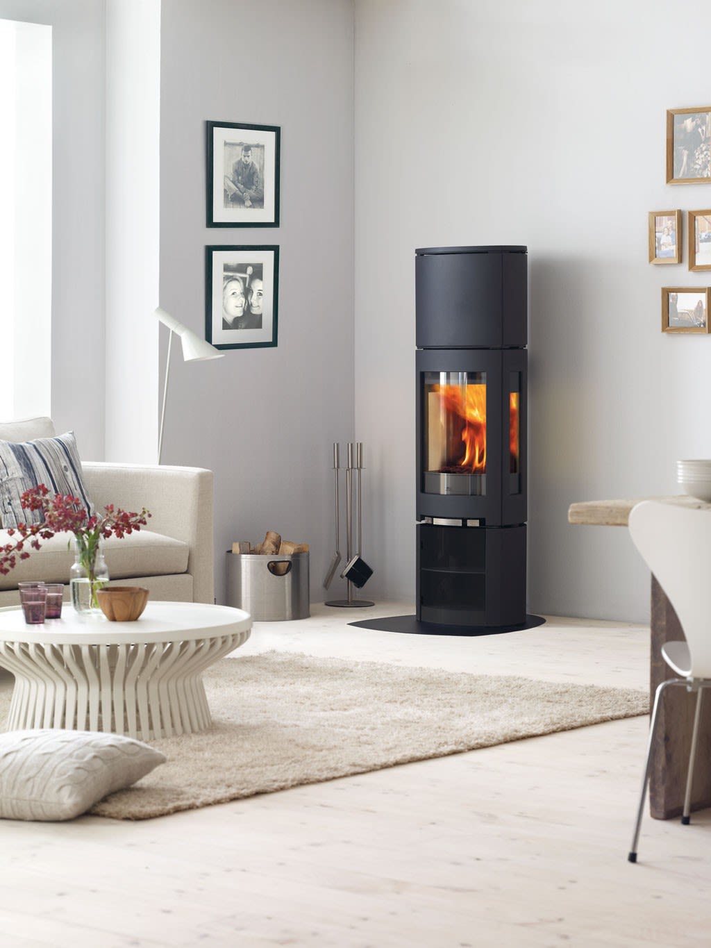 Lys stue med kremhvit sofa, rundt hvitt bord, beige teppe og svart vedovn med flammer.