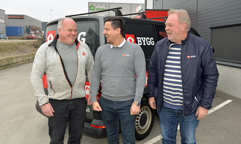 Tre menn i arbeidstøy foran svart varebil med rødt-hvitt BYGG-logo på parkeringsplass