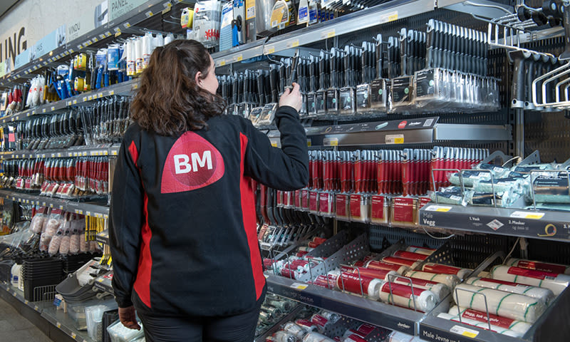 Ansatt i svart-rød BM-genser tar pensel fra hylle med pensler og hvite maleruller