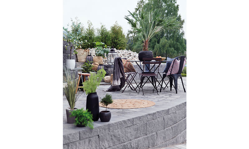 Uteplass med grå steinhelle, svart metallbord og stoler, grønne planter og naturfargede puter