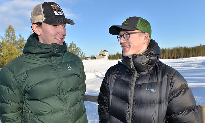 To menn i grønne og svarte dunjakker og caps ved snødekt gjerde foran hus og skog.