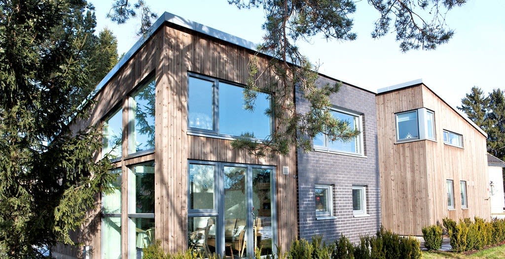 Moderne enebolig i lys tre- og grå mursteinsfasade med store vinduer, omgitt av furutrær