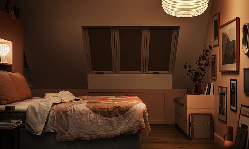 Soverom under skråtak med varm oransje belysning, seng ved takvinduer, bilder og planter.