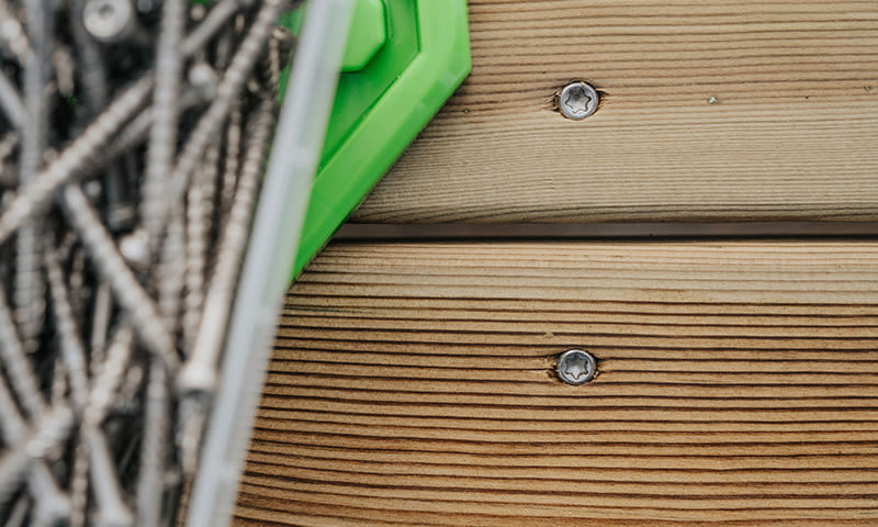 Nærbilde av grå skruer i hvit beholder med grønt lokk på lyst tredekke.