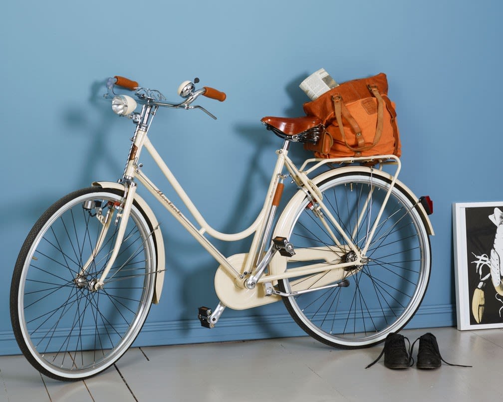 Kremfarget bysykkel med brunt sete og veske med avis mot blå vegg, svarte sko på gulvet.