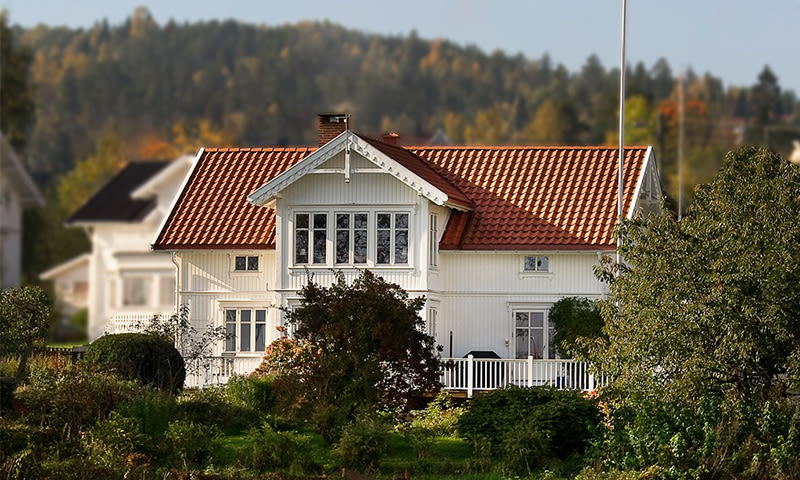 Hvit toetasjes trehus med rødt tegltak, store vinduer, hage og skog i bakgrunnen
