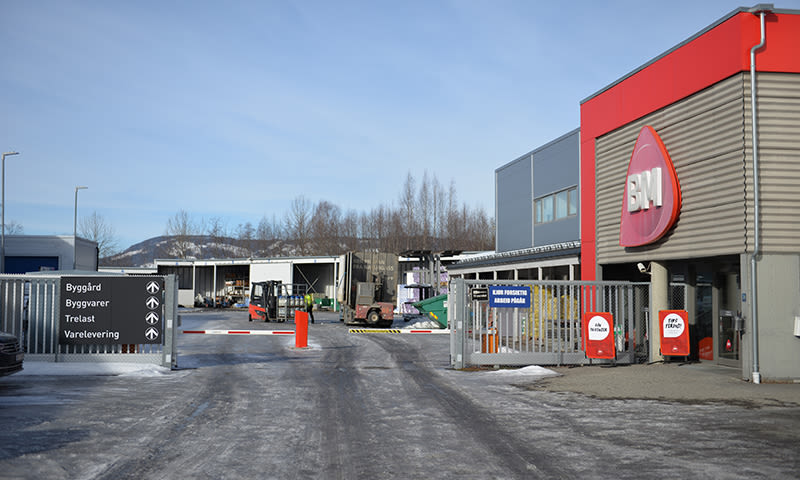 Åpen inngangsport til BM-byggvarehus med rødgrå fasade, gaffeltruck og blå himmel.