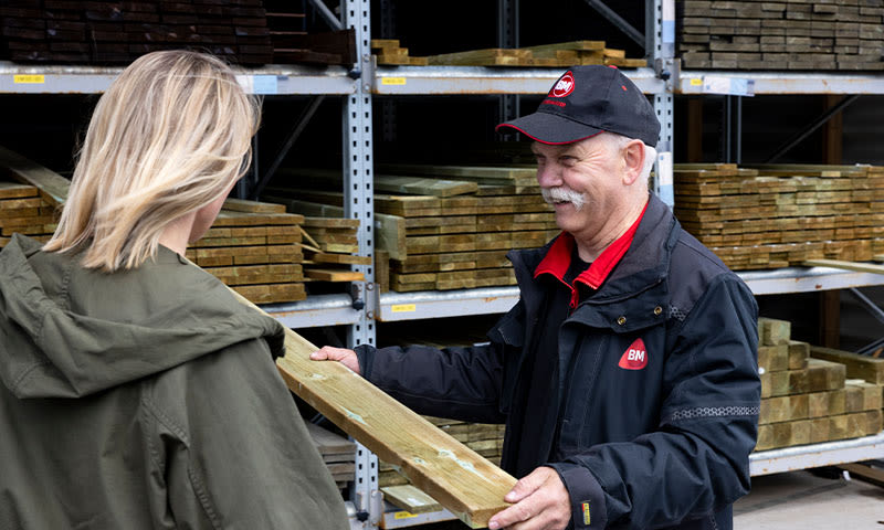 Ansatt i svart jakke gir trykkimpregnert treplanke til kvinne i grønn jakke foran hyller.