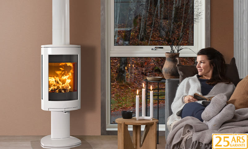 Hvit vedovn i stue, tente lys på trebord, grå sofa med pledd og bok, høstskog utenfor vindu