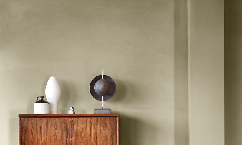 Lyst treskap foran olivengrønn vegg med hvit keramikkvase, liten krukke og metallskulptur.