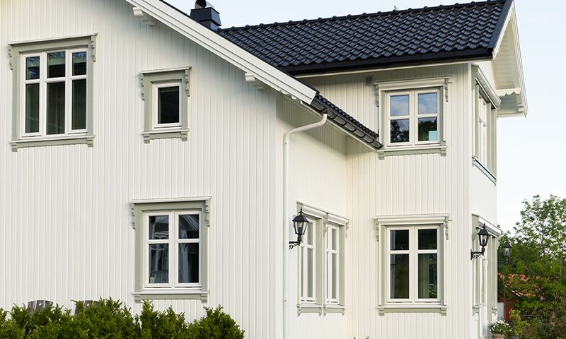 Hvit toetasjes trevilla med svart tak, hvite vinduer, utelamper og grønn hekk.